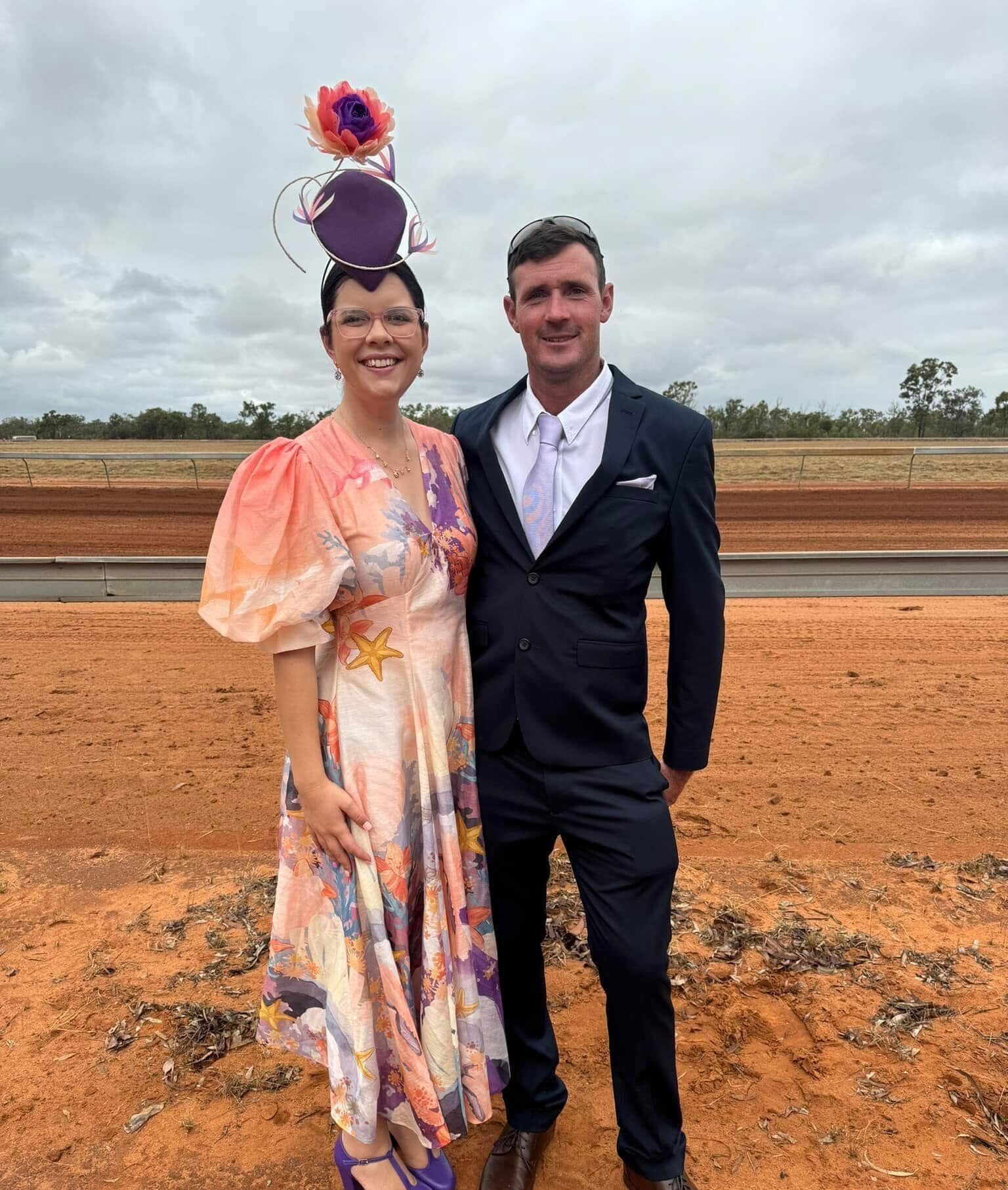 Smiling couple in race day attire stands on red dirt; woman wears a floral dress and purple headpiece with a rose.