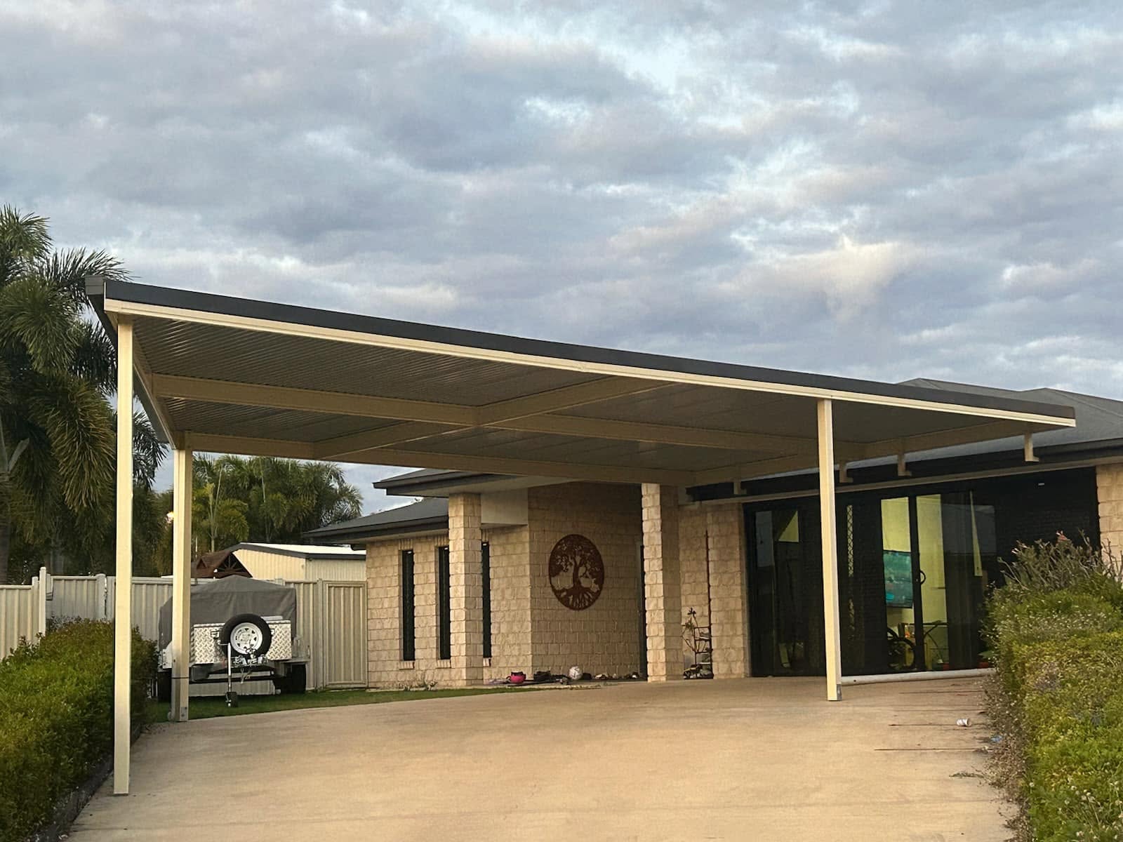 Beige flat-roof carport with cream steel posts extending over a concrete driveway in front of a light brick home under a dramatic cloudy sky.