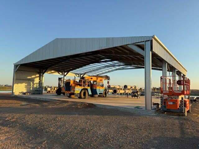 Large open steel-framed shed with corrugated roof housing orange construction equipment on gravel at sunset.