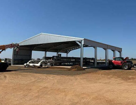 Open-front metal machinery shed with red tractor parked inside on dirt ground under clear blue sky